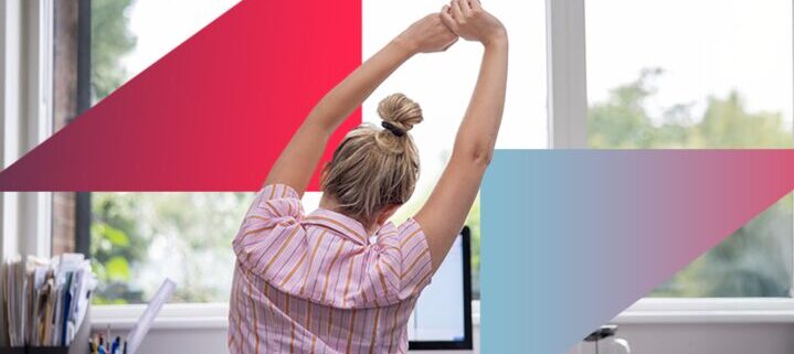 A person seated at a desk in front of a computer, viewed from behind, stretching their arms overhead with fingers interlaced.