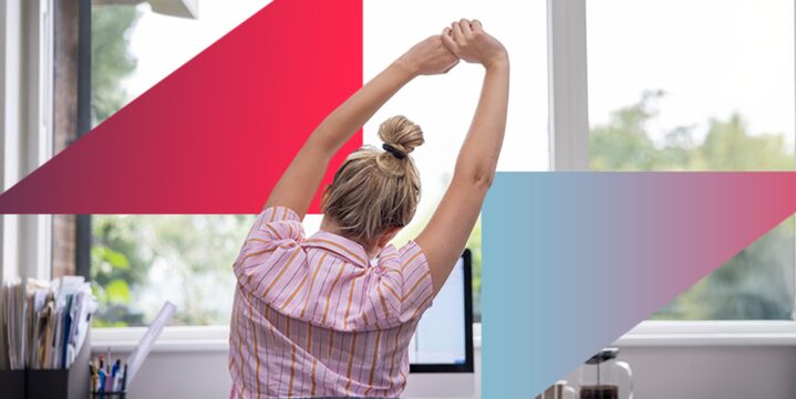 A person seated at a desk in front of a computer, viewed from behind, stretching their arms overhead with fingers interlaced.